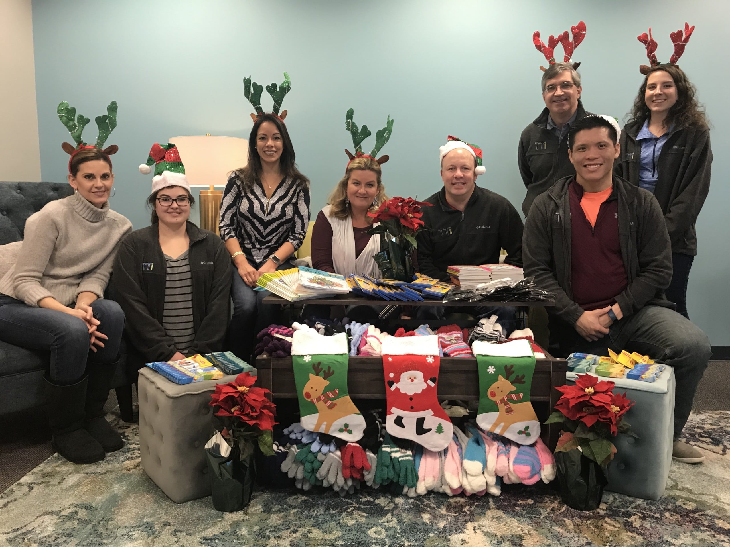 Join Our Team 1 TTi's team smiling with a collection of gifts collected for NESAP including Christmas stockings, mittens, books, and other items. The team are wearing Christmas hats & reindeer antlers.